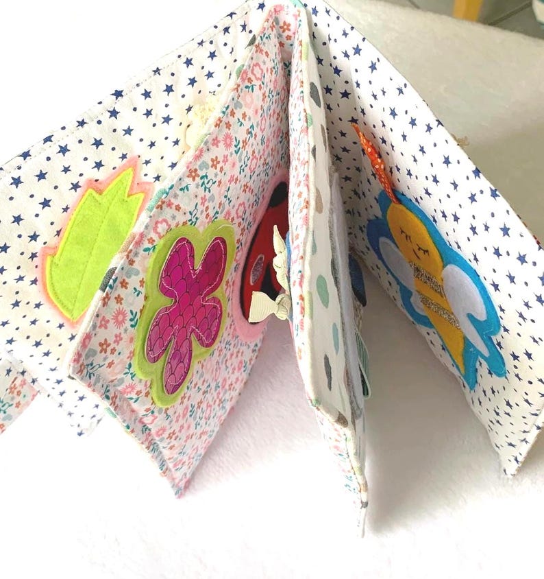 May include: A fabric baby book with appliqu&eacute;d designs. The pages display floral and star patterns, with felt shapes of a leaf, a flower, a ladybug, and a bee. The book is designed to stimulate a baby's senses with its textures and bright colours.