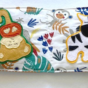 May include: A white fabric baby book with colorful embroidered animals. The book features a monkey, a zebra, and other jungle animals.