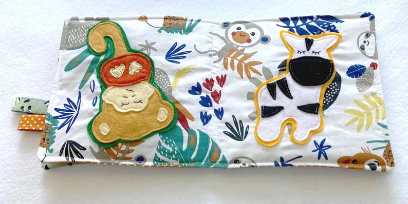 May include: A white fabric baby book with colorful embroidered animals. The book features a monkey, a zebra, and other jungle animals.