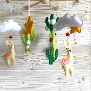 May include: A baby mobile with a desert theme. It features two llamas, two cacti, two clouds, two stars, and colorful beads. The mobile is made of felt and is hung from a wooden frame.