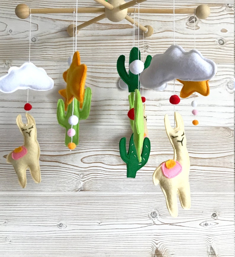 May include: A baby mobile with a desert theme. It features two llamas, two cacti, two clouds, two stars, and colorful beads. The mobile is made of felt and is hung from a wooden frame.