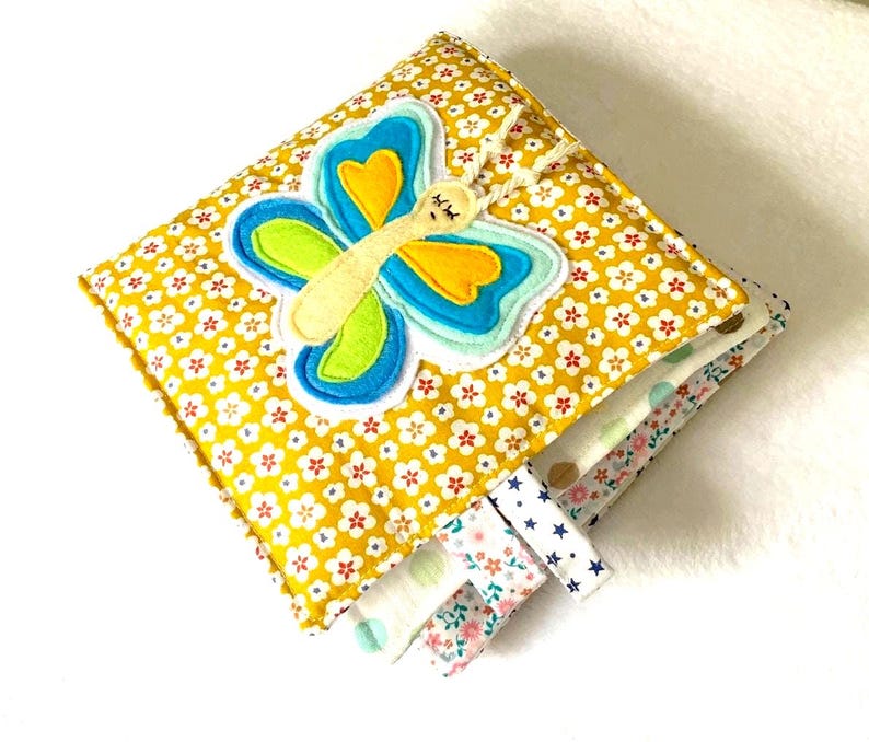 May include: A handmade, square baby book with a yellow floral pattern and a felt butterfly applique. The butterfly has blue, green, and yellow wings. Colourful fabric tags extend from the side, offering sensory stimulation for infants.