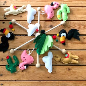 May include: A wooden mobile with felt animal and plant decorations. The mobile features a toucan, llama, flamingo, cactus, cloud, and a green leaf.