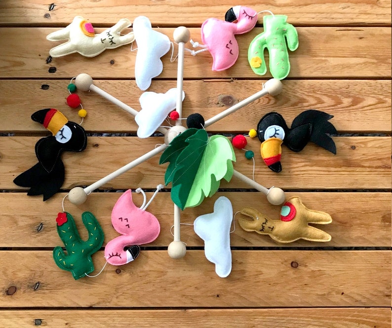 May include: A wooden mobile with felt animal and plant decorations. The mobile features a toucan, llama, flamingo, cactus, cloud, and a green leaf.