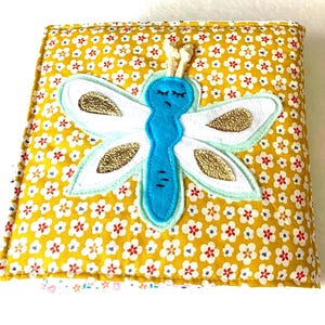 May include: A handmade fabric book with a yellow floral pattern. A blue dragonfly with white wings and gold accents is appliqu&eacute;d on the cover. The book is closed with a cream-coloured cord.
