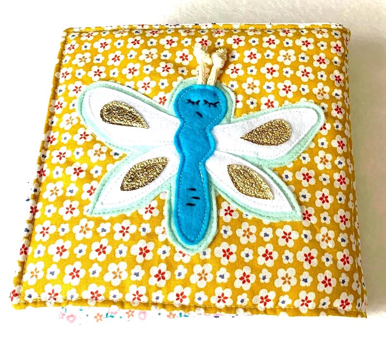 May include: A handmade fabric book with a yellow floral pattern. A blue dragonfly with white wings and gold accents is appliqu&eacute;d on the cover. The book is closed with a cream-coloured cord.