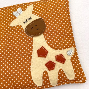 May include: A beige felt giraffe with brown felt spots and a sparkly silver tail is sewn onto a brown fabric with white polka dots.