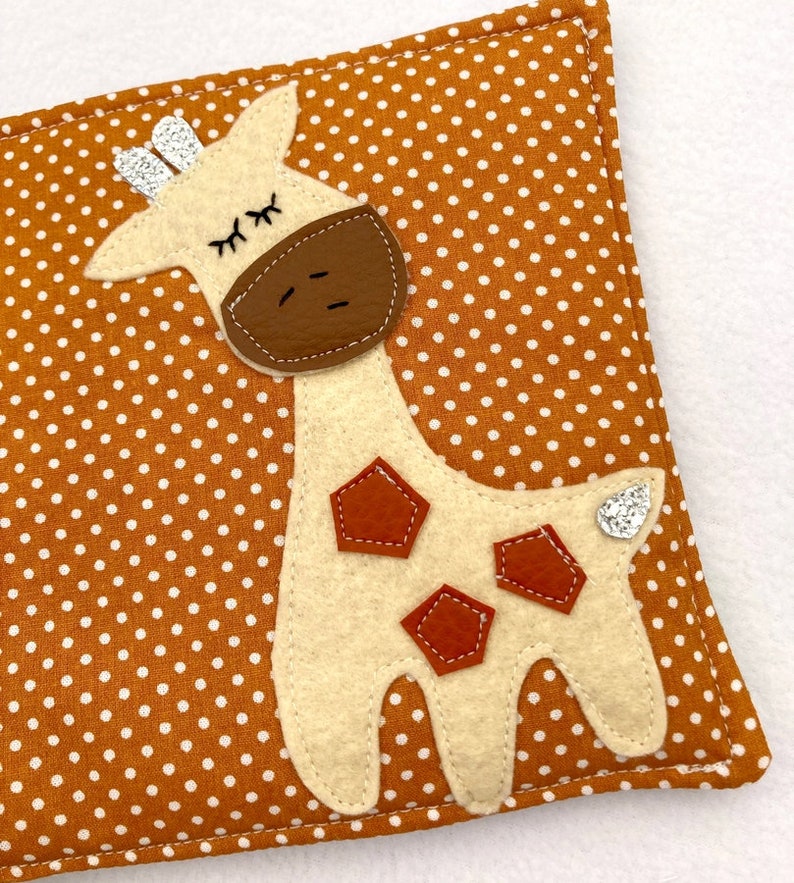 May include: A beige felt giraffe with brown felt spots and a sparkly silver tail is sewn onto a brown fabric with white polka dots.