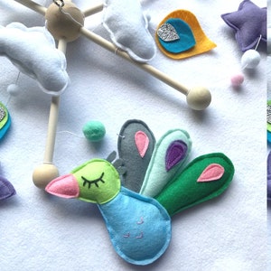 May include: A baby mobile with a green, blue, and gray peacock, white clouds, a yellow and blue teardrop, a purple star, and white and pink pom-poms.