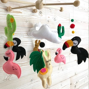 May include: A colorful felt baby mobile with a white cloud, two pink flamingos, a yellow llama, a green palm leaf, two black toucans, and two green cacti. The mobile is hanging from a wooden frame.