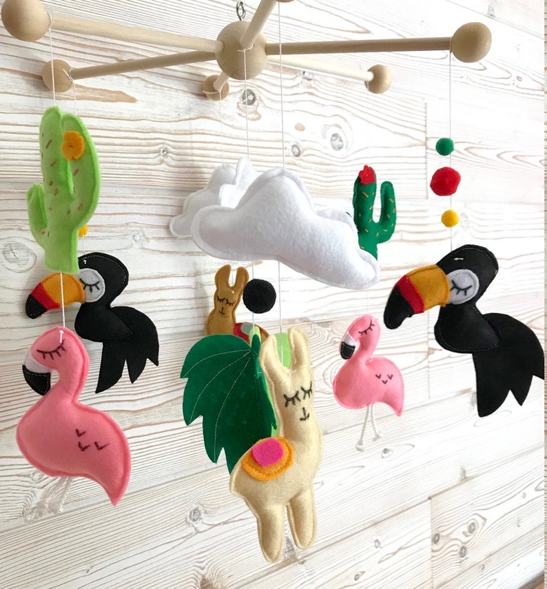 May include: A colorful felt baby mobile with a white cloud, two pink flamingos, a yellow llama, a green palm leaf, two black toucans, and two green cacti. The mobile is hanging from a wooden frame.