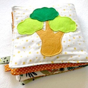May include: A white fabric book with a yellow tree and green leaves. The book has a white background with yellow polka dots. The book is made of soft fabric and is perfect for babies and toddlers.
