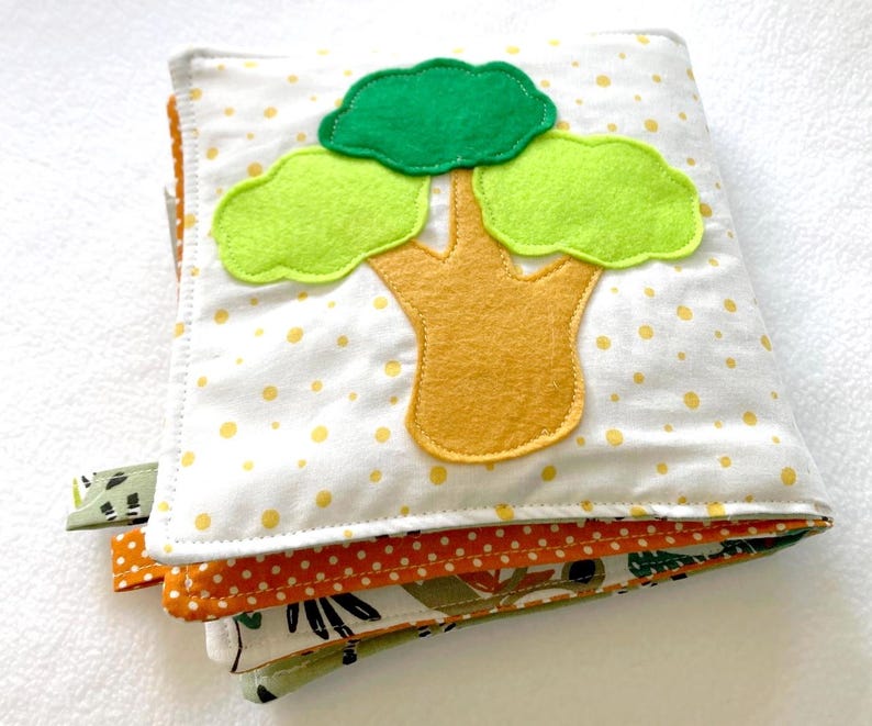 May include: A white fabric book with a yellow tree and green leaves. The book has a white background with yellow polka dots. The book is made of soft fabric and is perfect for babies and toddlers.