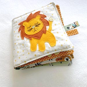 May include: A multi-page soft book for babies and toddlers featuring a yellow lion with a brown mane and a white background with yellow polka dots. The book has multiple pages with different animal designs.