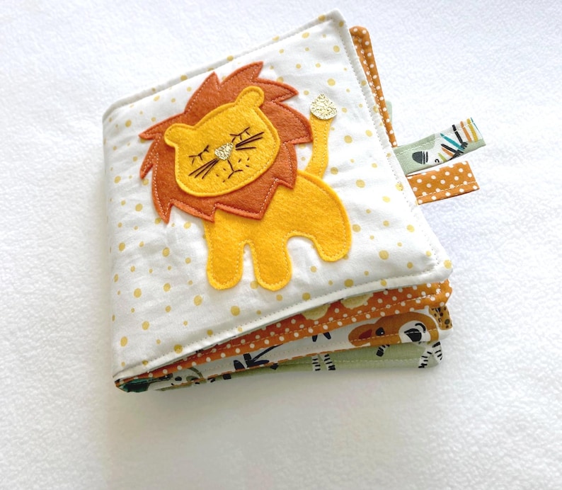 May include: A multi-page soft book for babies and toddlers featuring a yellow lion with a brown mane and a white background with yellow polka dots. The book has multiple pages with different animal designs.