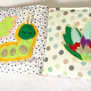 May include: Open cloth book with colourful appliqu&eacute;s. One page features a green caterpillar with gold accents and a leaf. The other page has a floral design with green, red, and purple accents. The background is white with star and polka dot patterns.