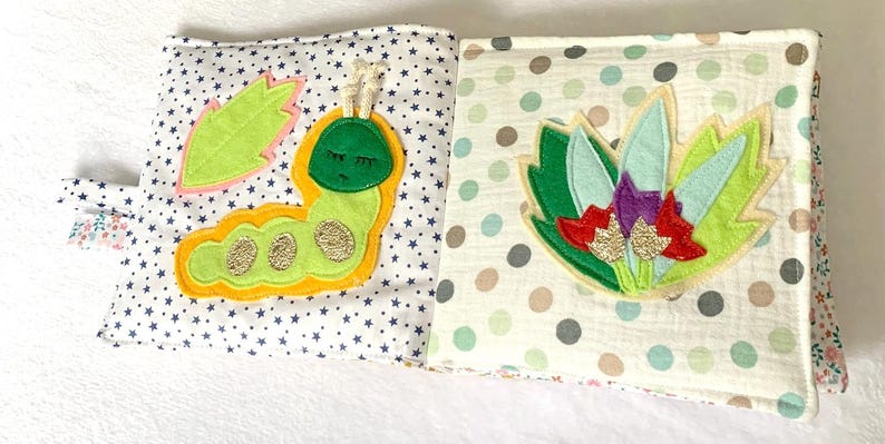 May include: Open cloth book with colourful appliqu&eacute;s. One page features a green caterpillar with gold accents and a leaf. The other page has a floral design with green, red, and purple accents. The background is white with star and polka dot patterns.