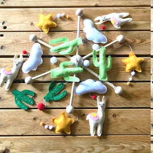 May include: A wooden mobile with felt llama, cactus, star, and cloud shapes. The mobile is suspended from a wooden ring and has a white, light blue, green, yellow, and pink color scheme.