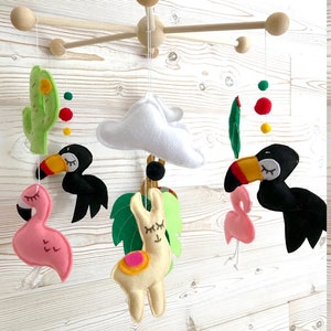 May include: A wooden baby mobile with felt animal toys. The mobile features a llama, two flamingos, two toucans, a cactus, and a cloud. The mobile is decorated with colorful felt balls.