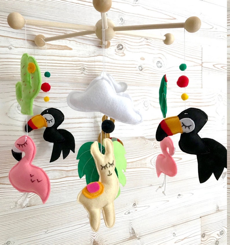 May include: A wooden baby mobile with felt animal toys. The mobile features a llama, two flamingos, two toucans, a cactus, and a cloud. The mobile is decorated with colorful felt balls.