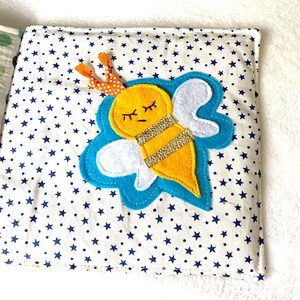 May include: A fabric book page with a yellow felt bee, gold glitter stripes, white wings, and closed eyes. The bee is on a blue cloud shape, set against a white background with blue star patterns. The bee has orange and white polka dot antennae.