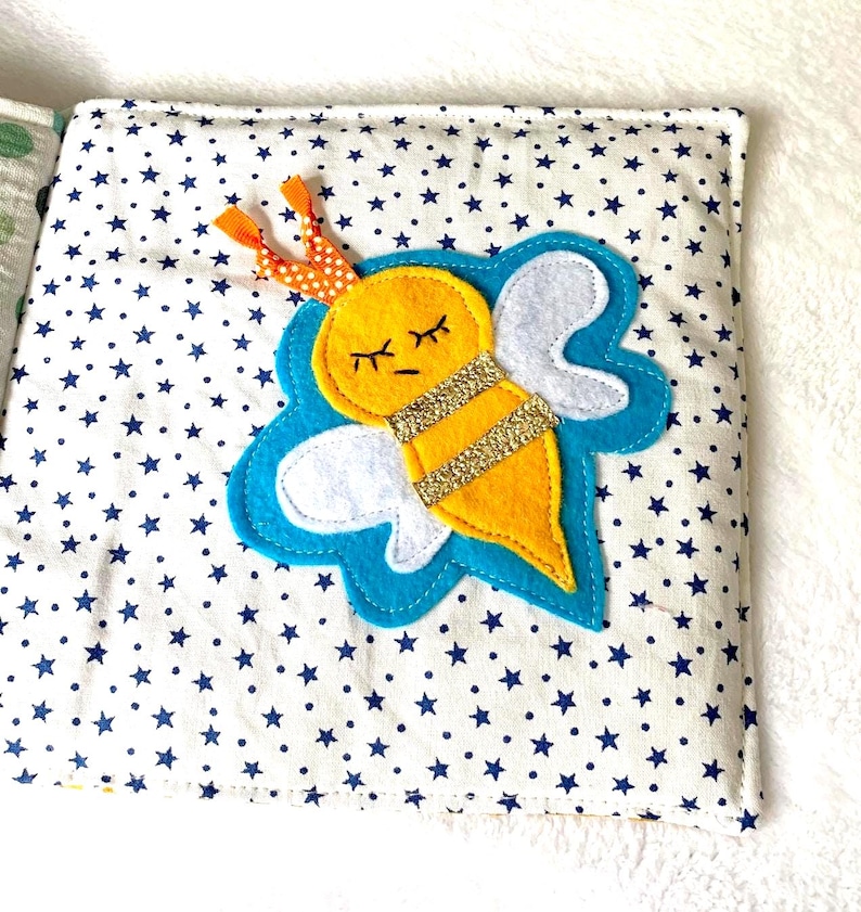 May include: A fabric book page with a yellow felt bee, gold glitter stripes, white wings, and closed eyes. The bee is on a blue cloud shape, set against a white background with blue star patterns. The bee has orange and white polka dot antennae.
