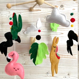 May include: A wooden baby mobile with felt animal and plant decorations. The mobile features a pink flamingo, a brown llama, a green cactus, a green leaf, and two white clouds. There are also black, red, and yellow pom-poms.