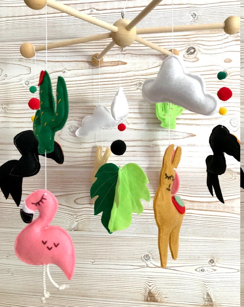 May include: A wooden baby mobile with felt animal and plant decorations. The mobile features a pink flamingo, a brown llama, a green cactus, a green leaf, and two white clouds. There are also black, red, and yellow pom-poms.