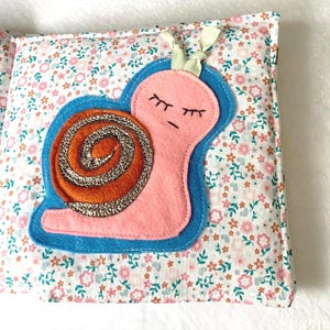 May include: A fabric book page with a pink snail and glittery orange shell, outlined in blue. The background is a floral pattern in pink, white, and green. A cream ribbon is attached at the top of the page.
