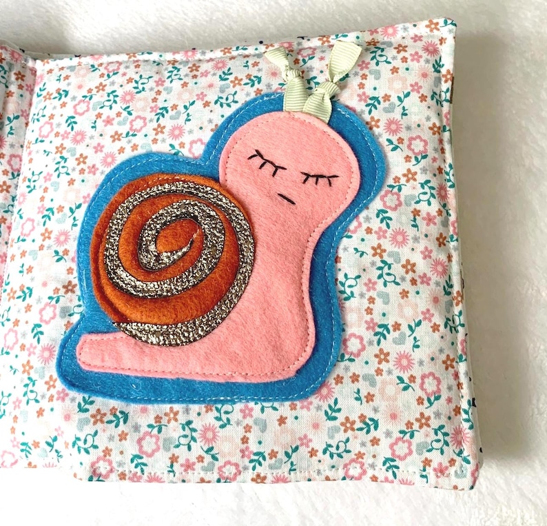 May include: A fabric book page with a pink snail and glittery orange shell, outlined in blue. The background is a floral pattern in pink, white, and green. A cream ribbon is attached at the top of the page.