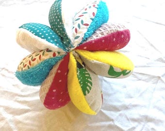 May include: A colorful, soft fabric flower toy with multiple petals. Each petal features a different pattern and color, including polka dots, floral prints, and solid colors like turquoise, pink, and yellow. The toy is designed for babies and young children.