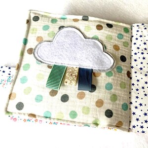 May include: A handmade baby book with a cloud design. The cover features a white cloud with grey trim and three colourful ribbons. The book has a polka dot pattern in green, brown, and grey, and a star pattern on the inside pages.