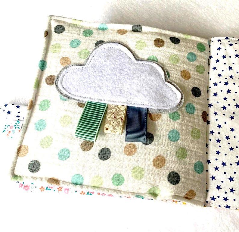 May include: A handmade baby book with a cloud design. The cover features a white cloud with grey trim and three colourful ribbons. The book has a polka dot pattern in green, brown, and grey, and a star pattern on the inside pages.