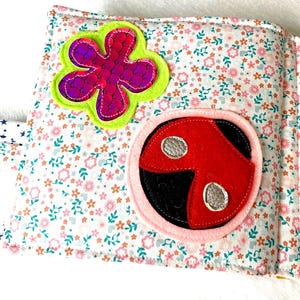 May include: An open fabric book with floral print pages. The pages display felt appliqu&eacute;s of a purple flower with a green border and a red ladybird with silver spots, set against a white background.