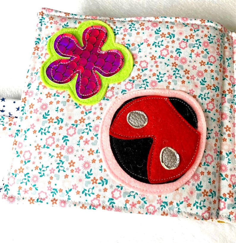 May include: An open fabric book with floral print pages. The pages display felt appliqu&eacute;s of a purple flower with a green border and a red ladybird with silver spots, set against a white background.