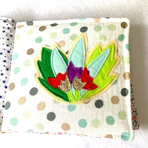 May include: A handmade fabric book with a colourful floral design. The cover features a felt flower with green, light blue, red, and gold glitter accents, set against a white polka dot background. The book's pages are visible, showcasing various patterns.