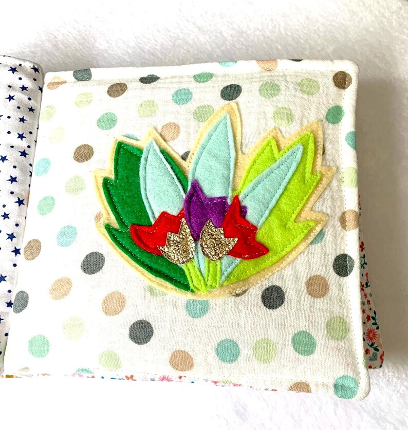 May include: A handmade fabric book with a colourful floral design. The cover features a felt flower with green, light blue, red, and gold glitter accents, set against a white polka dot background. The book's pages are visible, showcasing various patterns.