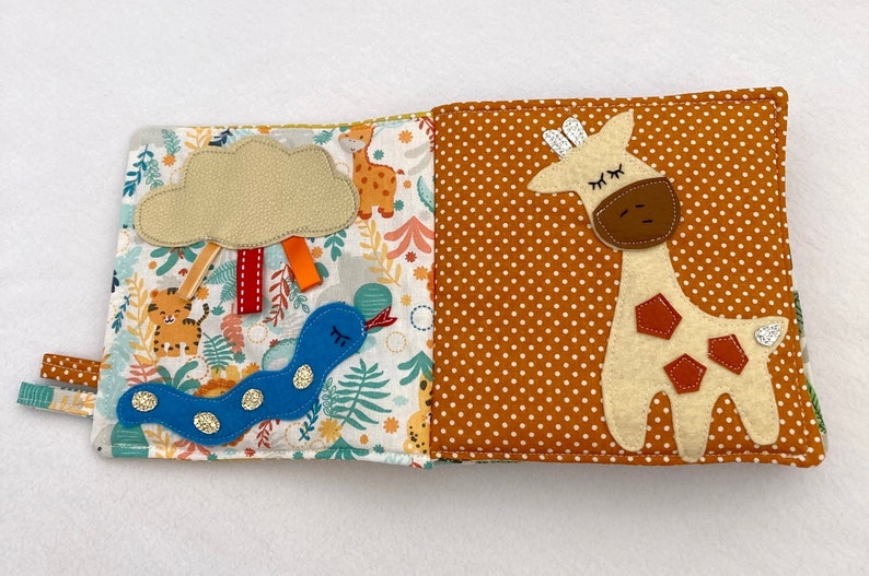 May include: A soft, multi-page book for babies and toddlers features a colorful jungle scene with a blue snake, a cloud, and a tiger. The opposite page features a brown and white giraffe with brown spots on a brown background with white polka dots.
