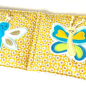 May include: A yellow fabric book with a floral pattern, featuring two appliqued butterflies. One butterfly is blue and white with gold glitter, the other is blue, yellow, and green. The book has a small tag with floral and star patterns.