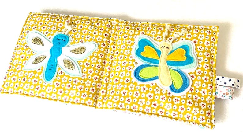 May include: A yellow fabric book with a floral pattern, featuring two appliqued butterflies. One butterfly is blue and white with gold glitter, the other is blue, yellow, and green. The book has a small tag with floral and star patterns.