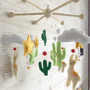 May include: A wooden baby mobile with felt cactus, llama, star, and cloud decorations. The mobile is suspended from a wooden frame with five arms.