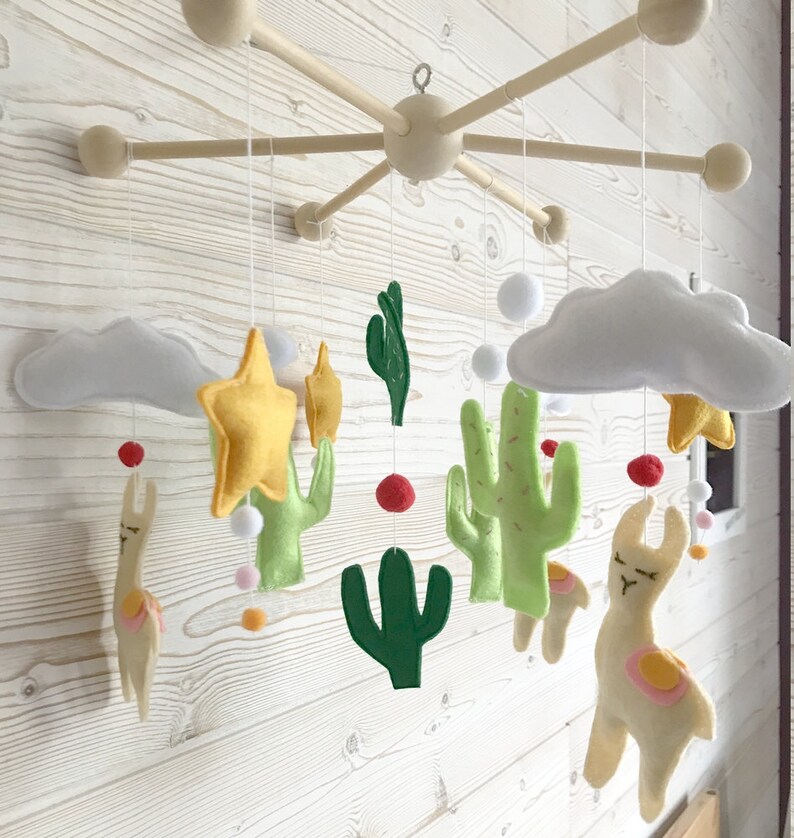 May include: A wooden baby mobile with felt cactus, llama, star, and cloud decorations. The mobile is suspended from a wooden frame with five arms.