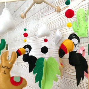 May include: A colorful felt baby mobile with a llama, toucans, clouds, a cactus, and a green leaf. The mobile is suspended from a wooden frame.