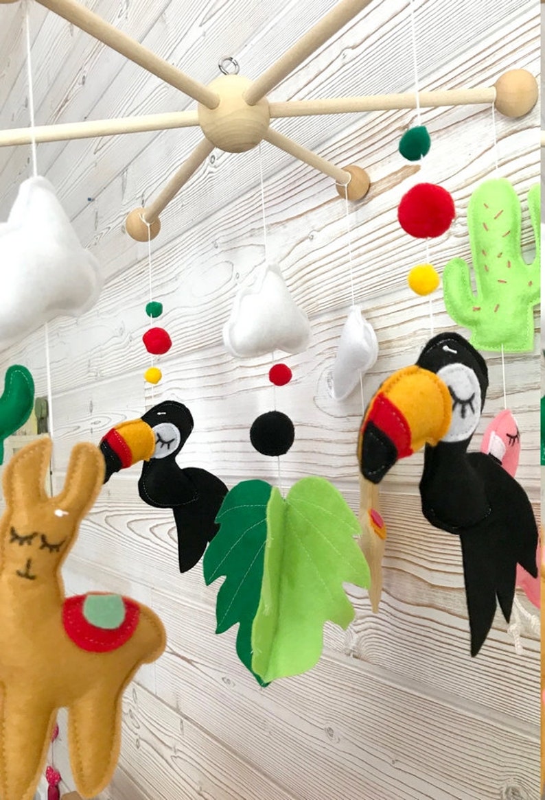 May include: A colorful felt baby mobile with a llama, toucans, clouds, a cactus, and a green leaf. The mobile is suspended from a wooden frame.