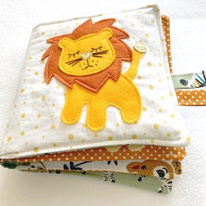May include: A multi-page baby sensory book with a yellow lion applique on the front cover. The book has a white background with yellow polka dots and a brown, yellow, and green patterned fabric on the inside pages.