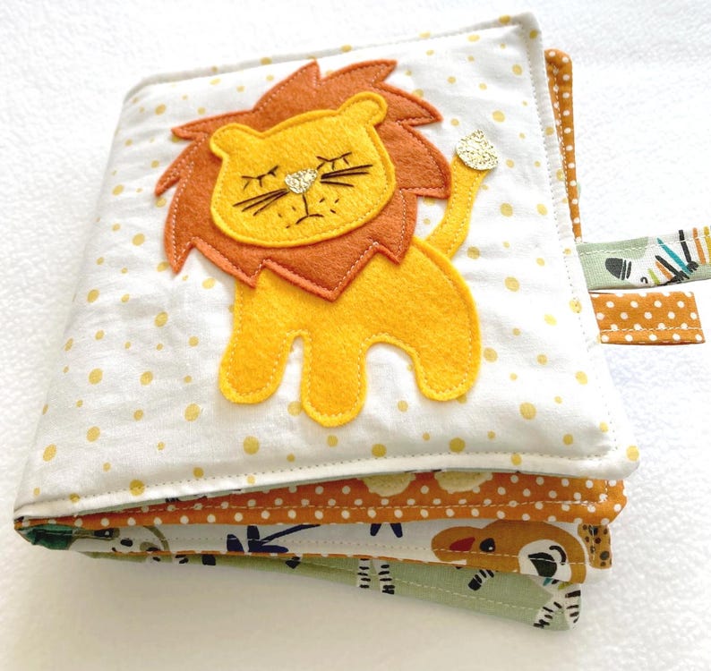May include: A multi-page baby sensory book with a yellow lion applique on the front cover. The book has a white background with yellow polka dots and a brown, yellow, and green patterned fabric on the inside pages.