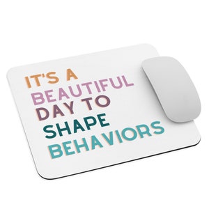 May include: White mousepad with the text "IT'S A BEAUTIFUL DAY TO SHAPE BEHAVIORS" in orange, purple, teal, and blue. A white computer mouse is positioned to the right of the mousepad. The mousepad is rectangular.