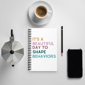 以下が含まれることがあります： 白い螺旋綴じのノートに、「It's a beautiful day to shape behaviors」というカラフルなテキストが書かれています。ノートは、白い表面にコーヒーカップ、鉛筆、黒いスマートフォンと一緒に置かれています。