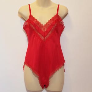 May include: A red bodysuit with thin straps and lace trim. The lingerie piece is displayed on a mannequin against a white background. The bodysuit has a V-neckline and delicate lace detailing along the edges.