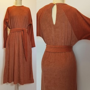 May include: A rust-colored dress with long sleeves and a matching belt. The dress features a round neckline and a keyhole detail at the back, fastened with a gold-colored button. The dress is displayed on a mannequin.